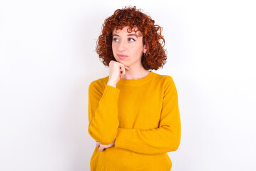young redhead girl wearing yellow sweater over white background with hand under chin and looking sideways with doubtful and skeptical expression, suspect and doubt.