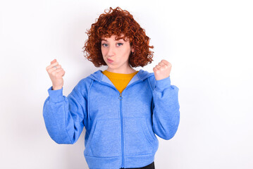 Irritated young redhead girl wearing blue jacket over white background blows cheeks with anger and raises clenched fists expresses rage and aggressive emotions. Furious model