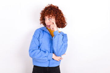 Astonished young redhead girl wearing blue jacket over white background looks aside surprisingly with opened mouth.