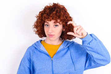 young redhead girl wearing blue jacket over white background purses lip and gestures with hand, shows something very little.