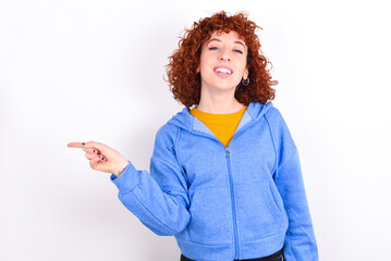 young redhead girl wearing blue jacket over white background laughs happily points away on blank space demonstrates shopping discount offer, excited by good news or unexpected sale.