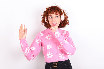 Happy young redhead girl wearing pink floral t-shirt over white background sings favourite song keeps hand near mouth as if microphone wears wireless headphones, listens music