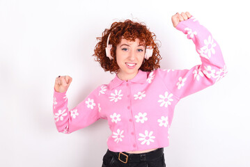 Carefree young redhead girl wearing pink floral t-shirt over white background with toothy smile raises arms dances carefree moves with rhythm of music listens music from playlist via headphones