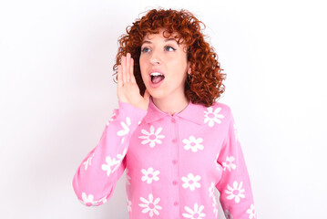 young redhead girl wearing pink floral t-shirt over white background look empty space holding hand near her face and screaming or calling someone.