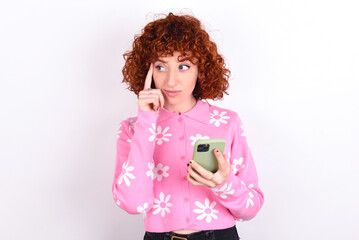 young redhead girl wearing pink floral t-shirt over white background  holding gadget while sticking out tongue
