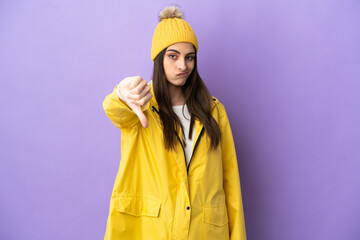Young caucasian woman wearing a rainproof coat isolated on purple background showing thumb down with negative expression
