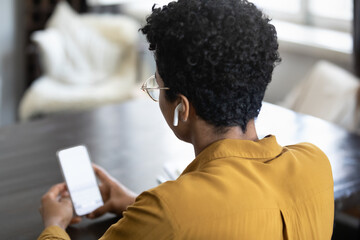 African American smartphone user woman with wireless earphone in ear holding mobile phone, sitting at table at home, talking on video call, attending online conference. Wireless connection concept