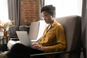 Serious millennial African student, homeowner woman, freelance employee working at laptop in armchair, using online app, virtual service, for studying, payment, browsing Internet