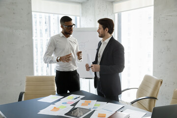 Focused young African American businessman in eyewear discussing working issues or paperwork with motivated Caucasian male colleague, standing at table in office, professional cooperation concept.