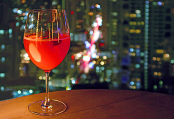 Glass of vibrant red cocktail on the rooftop terrace with blurry urban night view in background
