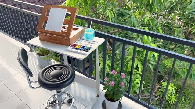 A beautiful home art studio on a balcony above green garden. Easel with watercolor paper and watercolour box on white table with cute chair and flower pot. Summer leisure.