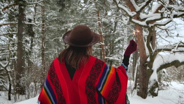 Happy positive hipster woman wearing in colorful ethnic wool poncho with sparklers in snowy forest. Person enjoy winter pleasures. Winter celebration and vacation season concept.