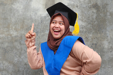 Happy graduate muslim girl wearing headscarf  with  a diploma, shows a gesture of victory and success, on a grey background. Indonesia Young Girl
