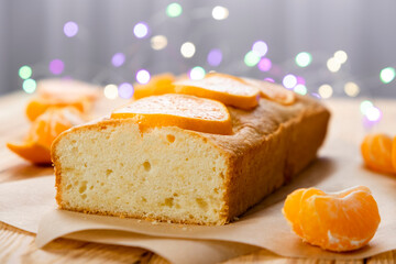 Loaf of delicious tangerine pound cake on parchment with pieces of mandarin and bokeh lights on rustic wooden background. Delicious breakfast, traditional tea time treat. Recipe of orange pie loaf.