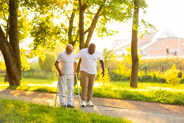 Caregiver is teaching old man to walk with walker. Professional nurse and patient walking outdoor at sunset. Assistance, rehabilitation and health care.