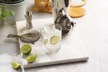 A glass with ice cubes, lime and refreshing beverage on marble board, shaker and bartender equipment