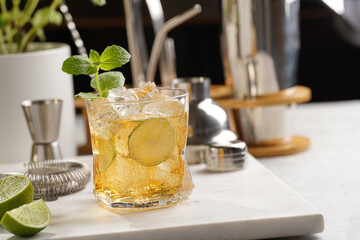 A glass with ice cubes, lime and refreshing orange beverage with mint on marble board, shaker and bartender equipment