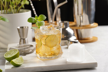 A glass with ice cubes, lime and refreshing orange beverage with mint on marble board, shaker and...