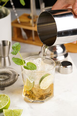 A glass with ice cubes, lime and refreshing orange beverage with mint on marble board, shaker and bartender equipment