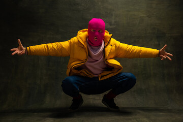 Fototapeta na wymiar Studio shot of young anonymous man wearing pink balaclava and yellow down jacket, coat isolated on dark vintage background. Concept of safety, art, fashion