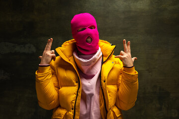 Studio shot of young anonymous man wearing pink balaclava and yellow down jacket, coat isolated on dark vintage background. Concept of safety, art, fashion - obrazy, fototapety, plakaty