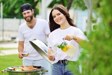 attractive bearded man fries meat in a barbecue, a young girl holds a glass jug with fresh lemonade