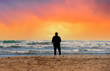 Man at beach on sunset during storm with waves in sea. Back side of European man 40-70s in jacket, standing and waiting or thinking about something, in feeling lonely sadness. Male at beach near sea.