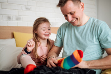 Father putting on different socks to his little daughter with Down syndrome when sitting on bed at...