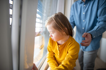 Sad little girl with Down syndrome looking throug window with her father at background.