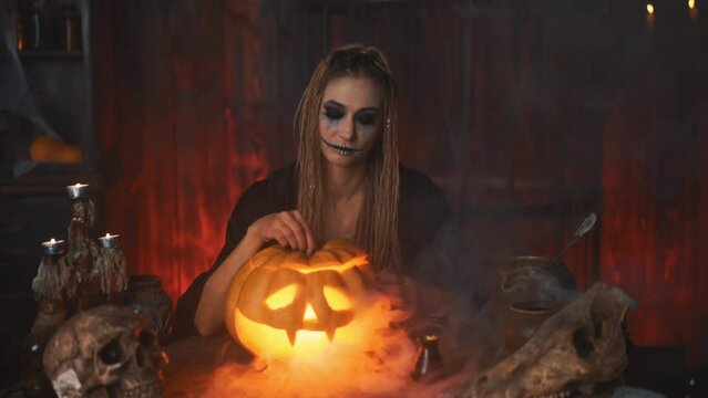 Come here. Halloween concept. Black witch Halloween pumpkin with carved smily face in hand sitting on the table dark room inviting with hand gesture. Jack o lantern head. Female necromancer