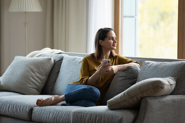 Young single woman sits on cozy sofa in living room holds cell phone smile looks out window, spend...