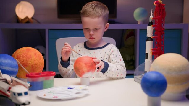 Blond boy paints planet solar system Mars with colorful paints sitting home table in evening, planets solar system, spaceships and space shuttle from constructor around. Cosmonautics Day on April 12.