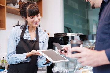 Young Caucasian barista woman is holding tablet for customer using smart phone scan QR code on...