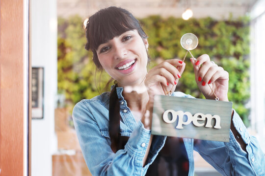 Smiling Caucasian Young barista woman or florist is wearing apron for setting open sign label on glass door in cafe or flowers shop. Start up for Coffee shop and cafe business Concept.