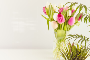 Easter home still life with tulips and decorative eggs in glass jar on a neutral grey background with home plants and copy space. Easter holiday composition with spring flower bouquet. Selective focus