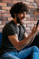 Vertical shot of happy excited young man looking at mobile phone screen, reading message with good news, celebrating victory and success, cheering emotion, gesturing with hand, at home.