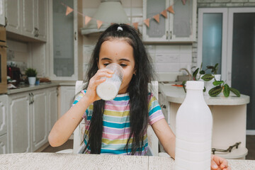 Cute little girl drinks milk at the table in the kitchen. the child is holding a bottle of milk....