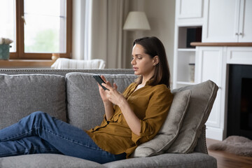 Side view young serious woman sit on sofa holds smartphone texting messages, share sms make...