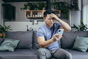 Man reading bad news from phone, asian frustrated and sad looking at phone screen sitting on sofa...