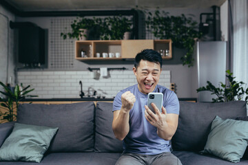 Obraz premium Happy Asian at home, won in the online uses the application on the smartphone, the man shows a gesture of victory, happy and satisfied sitting on the couch