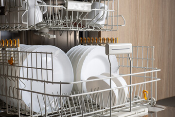 Open modern dishwasher with clean dishes. Pull-out shelves with white plates, mugs, spoons and...