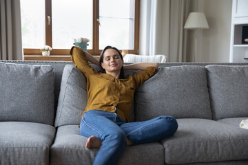 Obraz premium Serene young woman relaxing on sofa in cozy living room. Owner of modern apartments, stress-free leisure at home, climate control for comfort living, daydream, silent quiet domestic appliances concept