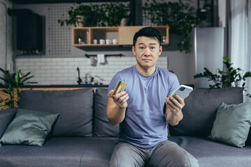 Fototapeta premium man alone at home, shopping online, asian uses phone and shopping app, holds credit card, sits on sofa at home