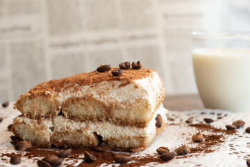 A piece of tiramisu with coffee beans on a lace napkin and a cup of milk on the table