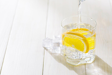 Pouring a glass of water with lemon and ice on a white table. Homemade lemonade with lemons slices