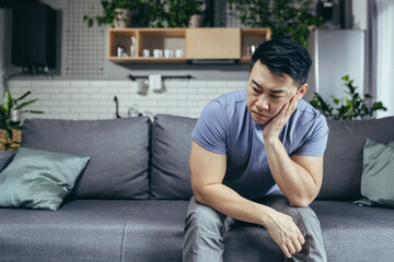 Upset Asian man at home, tired and depressed sitting on the couch in the living room