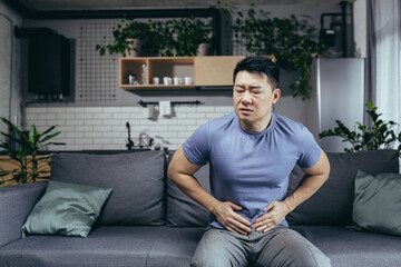Fototapeta premium Asian man himself at home sick sitting on the couch, has severe abdominal pain, man in the living room