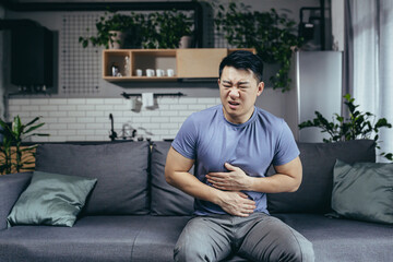 Asian man himself at home sick sitting on the couch, has severe abdominal pain, man in the living...