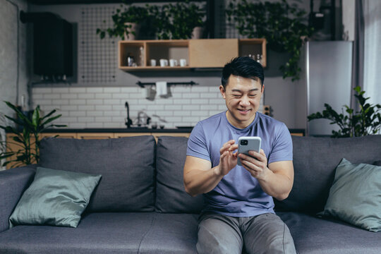 Happy asian at home sitting on sofa enjoying phone, typing message, cheerful and smiling man in loft living room