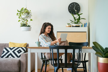 Cheerful smiling business woman talking on a video call meeting on her digital tablet while drink cup of coffee and sitting at home office desk